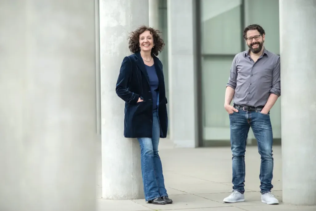 Two people are casually posing outside near large concrete pillars. The woman on the left is wearing a dark blazer and jeans, while the man on the right is in a gray shirt and jeans. Both are smiling and standing on a paved surface.