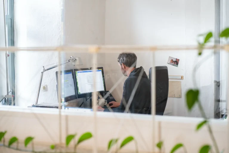 A person sits at a desk working on two computer monitors. They are holding a mug and appear focused. The scene is viewed through a glass screen with leaves partially visible in the foreground.