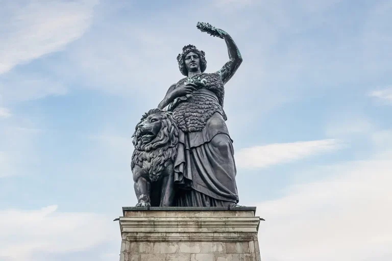 A large bronze statue depicting a woman in classical attire holding a wreath, symbolizing victory. She stands beside a lion on a pedestal against a backdrop of a lightly clouded sky.